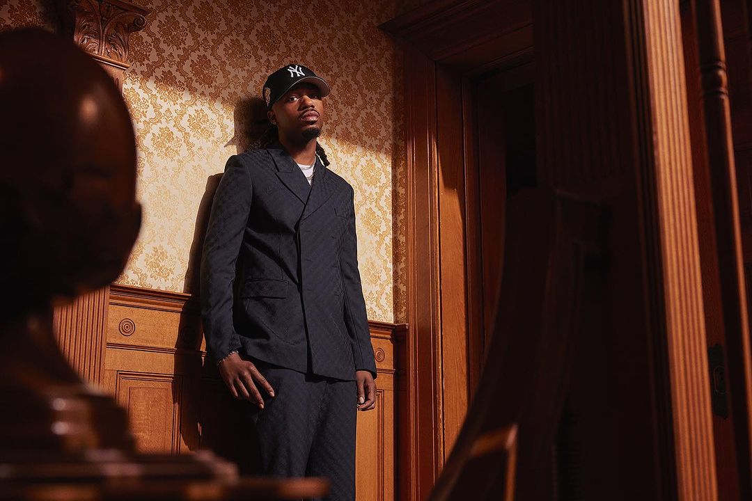 Adult man in a black suit and baseball cap standing indoors against a floral wallpaper background in a dimly lit home.