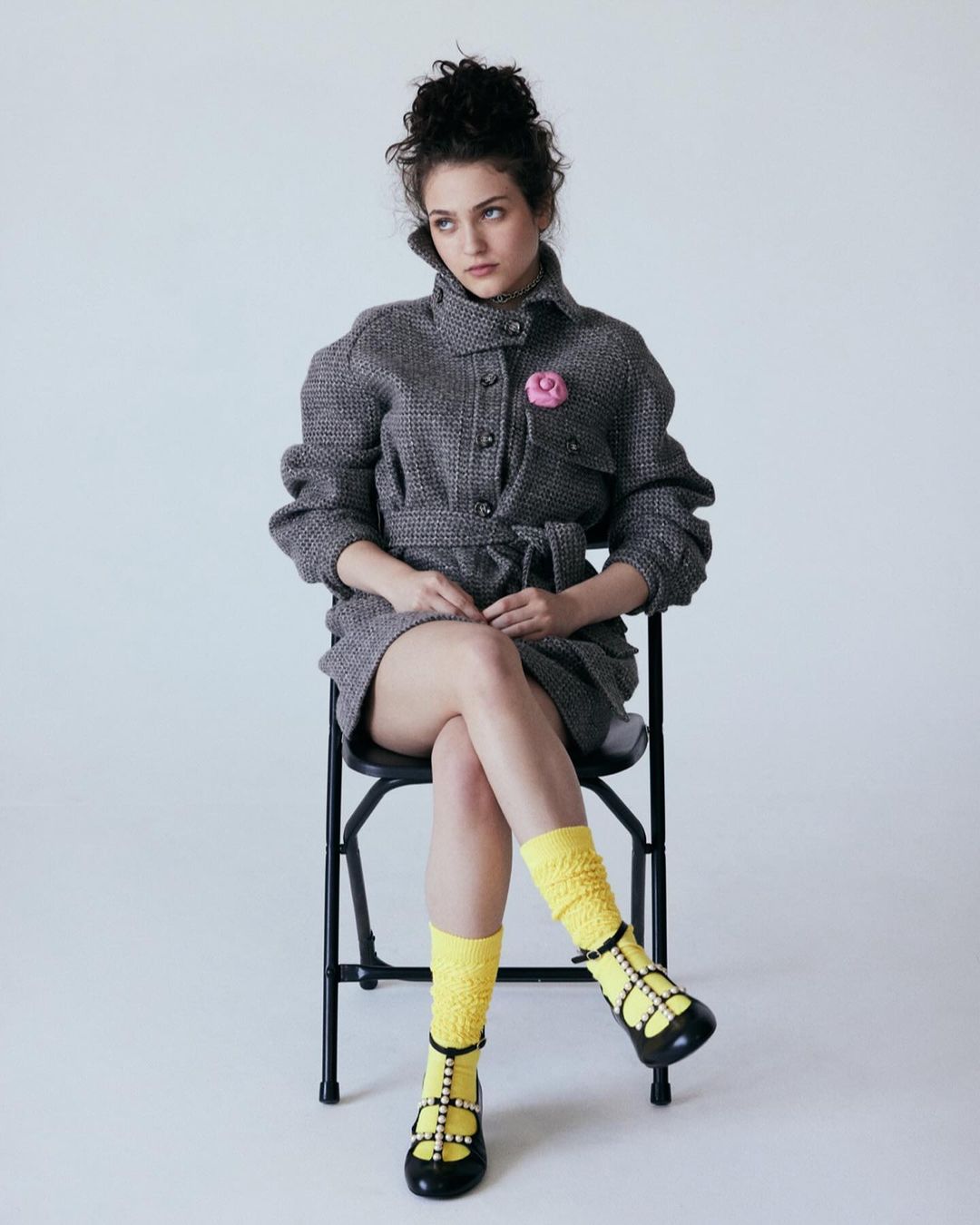 Young woman in tweed coat dress poses while seated in a photo studio. She wears yellow socks and black shoes, and has curly hair.