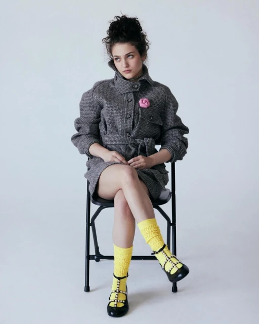 Young woman in tweed coat dress poses while seated in a photo studio. She wears yellow socks and black shoes, and has curly hair.