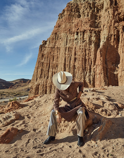 Person in cowboy hat and patterned robe sits on sandy hill with desert butte landscape background under a blue daytime sky.
