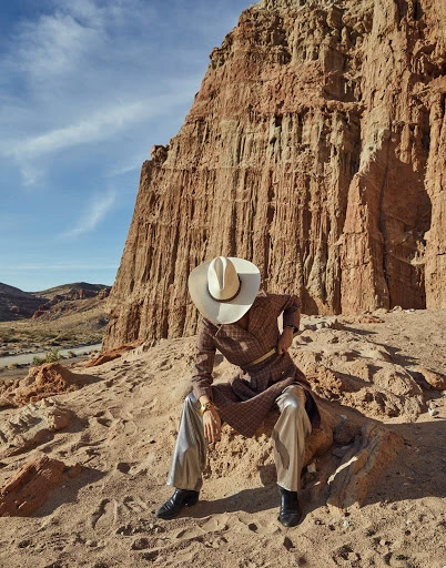 Person in cowboy hat and patterned robe sits on sandy hill with desert butte landscape background under a blue daytime sky.