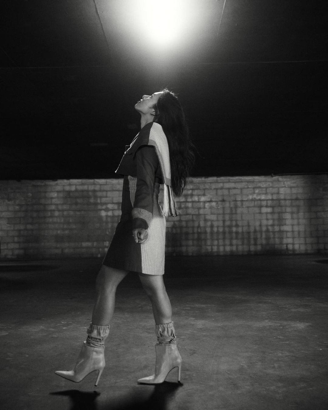 A woman in a two-tone dress and ankle boots confidently walks toward the viewer in a low-light urban environment with a brick wall.