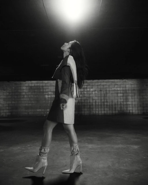 A woman in a two-tone dress and ankle boots confidently walks toward the viewer in a low-light urban environment with a brick wall.