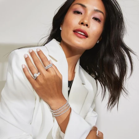 Smiling Asian woman wears diamond rings, bracelet, necklace, and earrings, accessorizing with a white blazer in a studio setting.