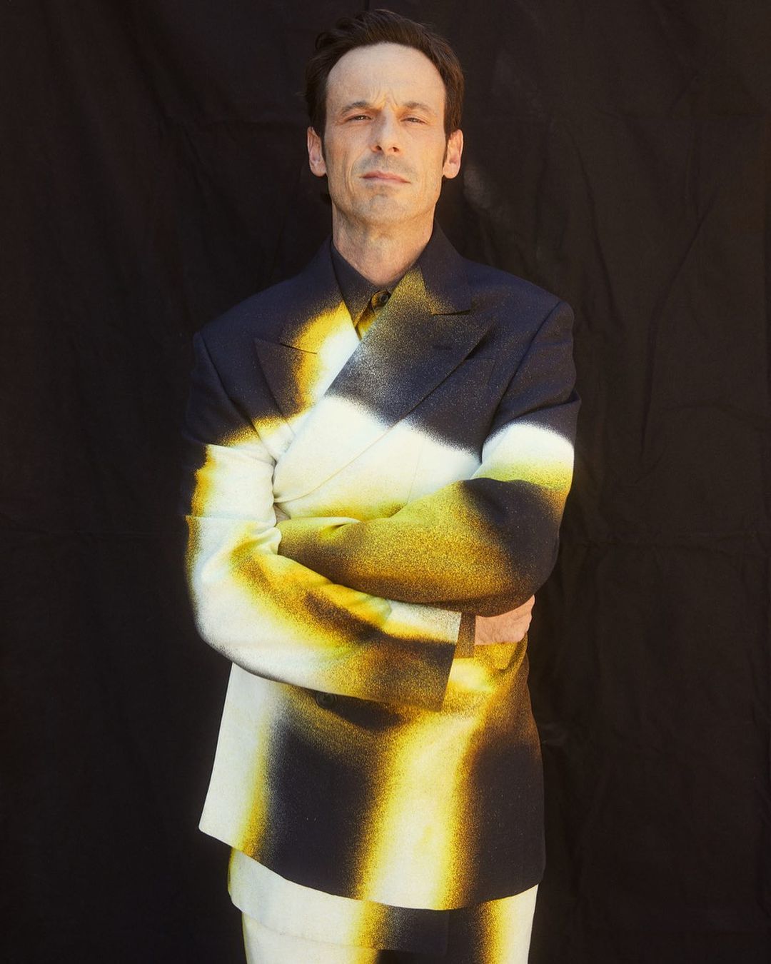 Man wearing a spray-painted double-breasted suit with arms crossed against a dark background, photographed by Sami Drasin.