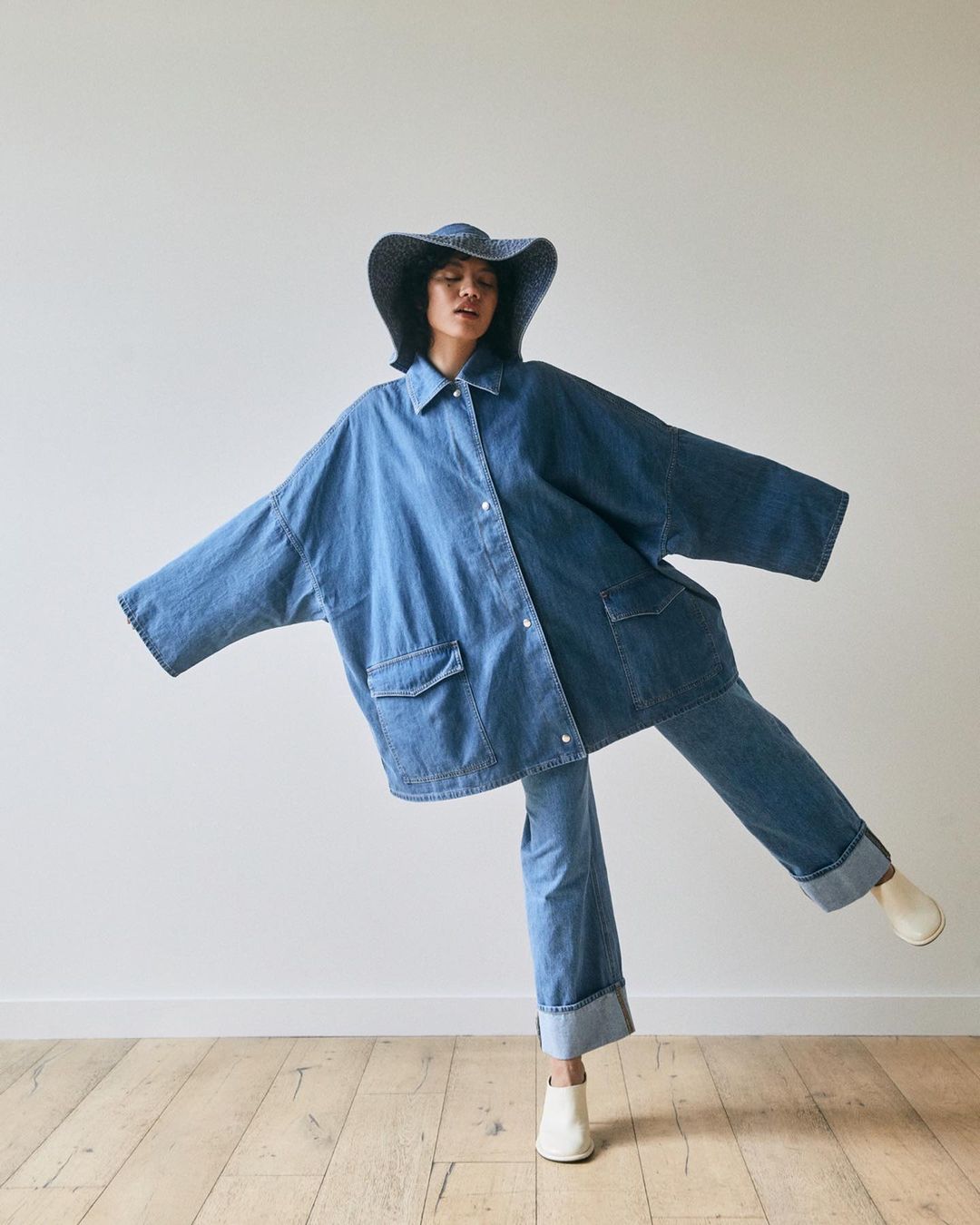 Woman in a denim outfit, including an oversized denim jacket and wide-brim hat, strikes a playful pose against a minimalist background.