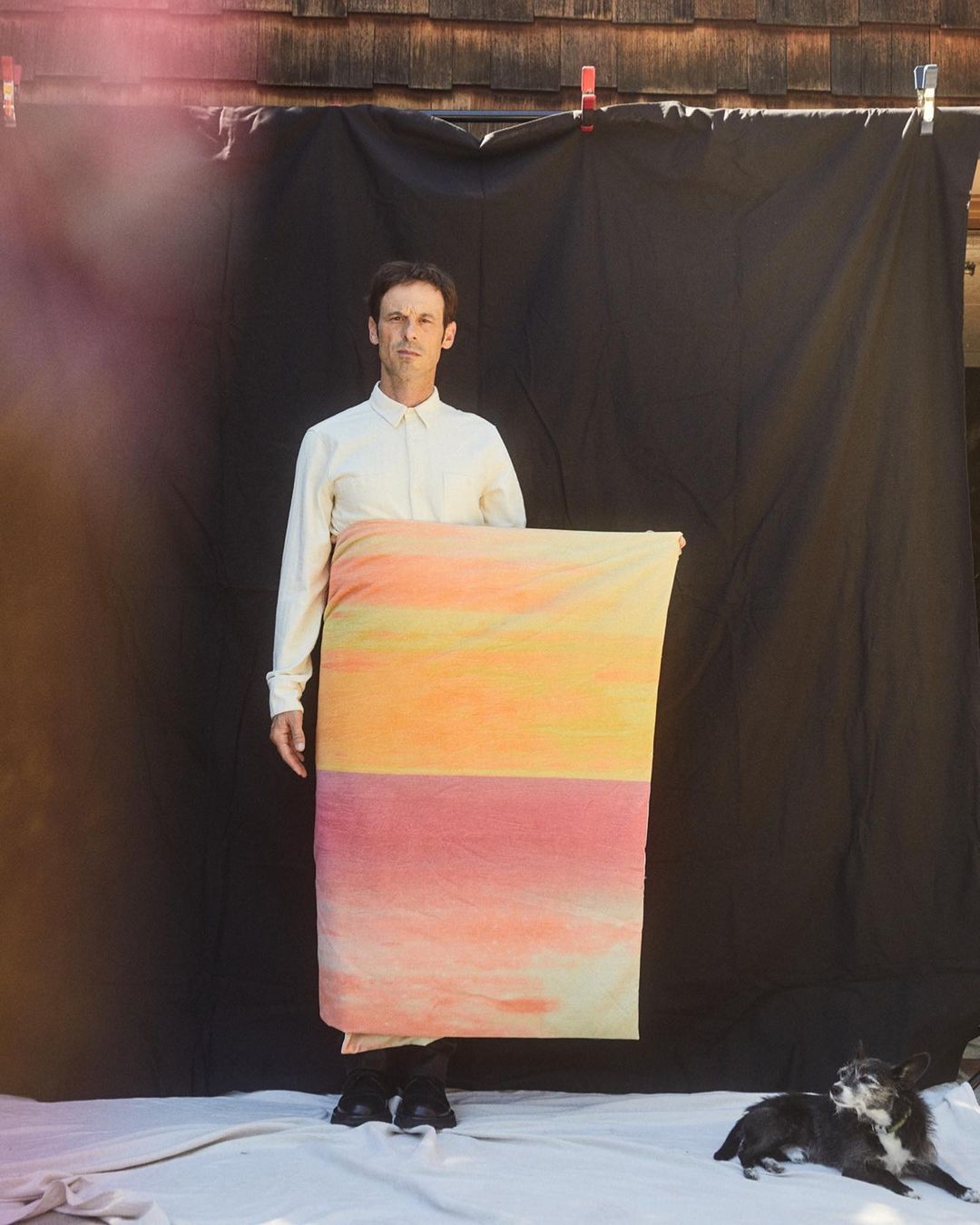 Adult male stands holding colorful fabric, black backdrop, outdoor setting with small black dog lying on white fabric.