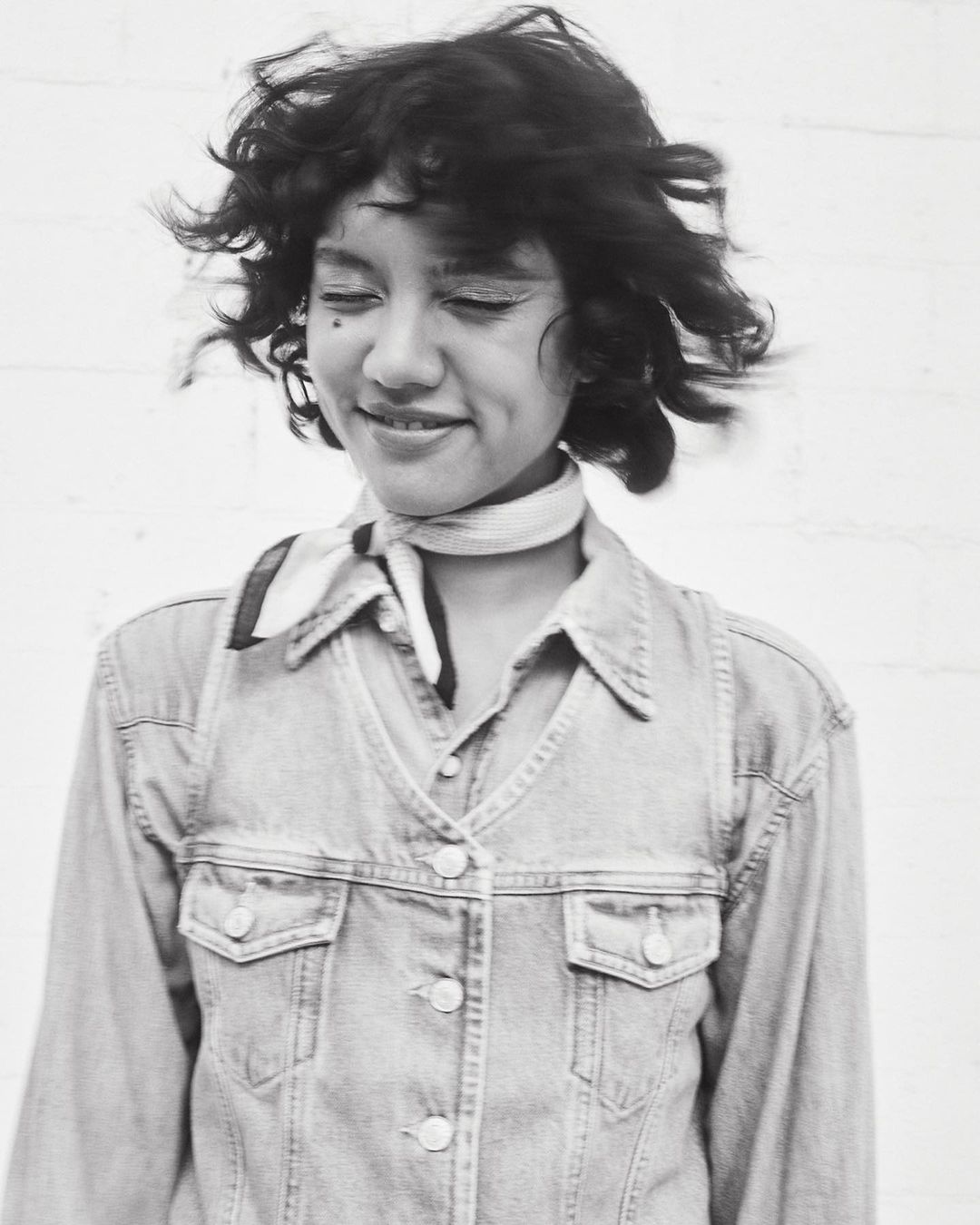 Portrait of a smiling woman with curly hair and a patterned neck scarf, wearing a denim jacket, in front of a white background.