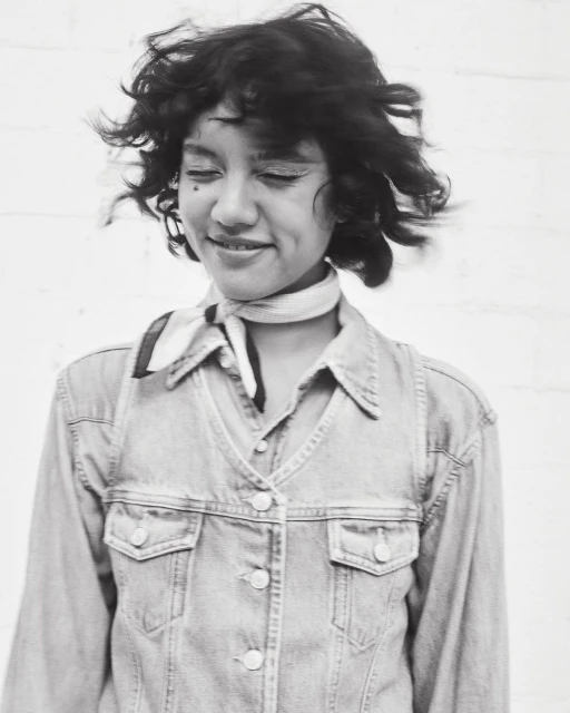 Portrait of a smiling woman with curly hair and a patterned neck scarf, wearing a denim jacket, in front of a white background.