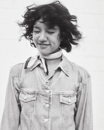 Portrait of a smiling woman with curly hair and a patterned neck scarf, wearing a denim jacket, in front of a white background.
