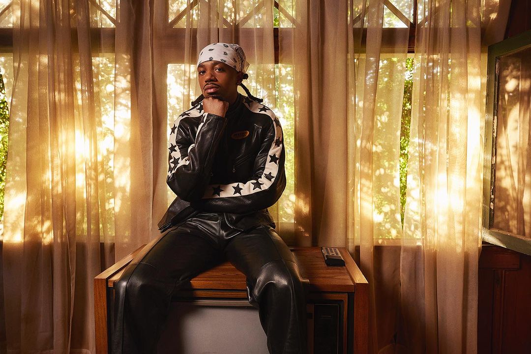 Adult male in bandana and leather jacket poses indoors sitting on a vintage television set in front of sheer curtains.