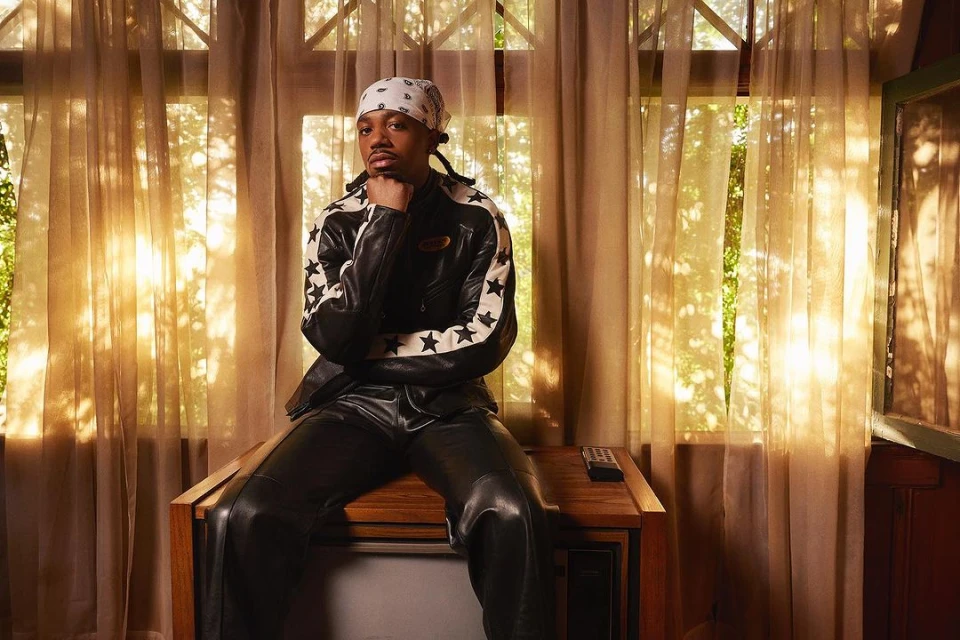 Adult male in bandana and leather jacket poses indoors sitting on a vintage television set in front of sheer curtains.
