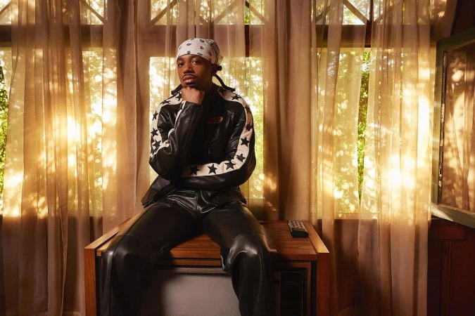 Adult male in bandana and leather jacket poses indoors sitting on a vintage television set in front of sheer curtains.