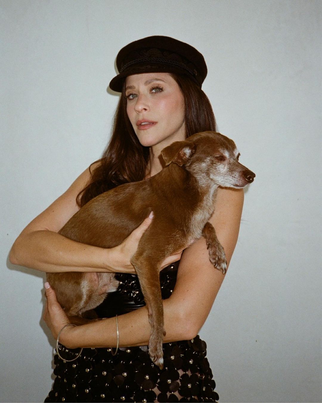 Woman wearing a black baker boy hat and dark dress holding a brown dog against a white background