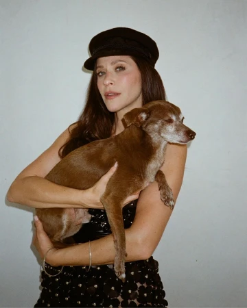 Woman wearing a black baker boy hat and dark dress holding a brown dog against a white background