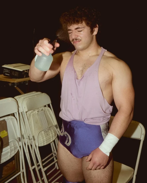 Professional wrestler with curly hair sprays water on his face in an indoor setting, wearing blue wrestling trunks and a sleeveless shirt.