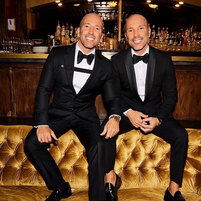 Two bald adult males in black tuxedos pose closely on a gold tufted couch in an indoor setting with a bar and bottles behind them.