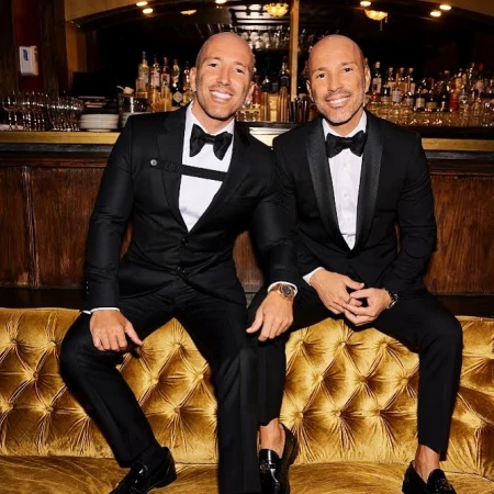 Two bald adult males in black tuxedos pose closely on a gold tufted couch in an indoor setting with a bar and bottles behind them.