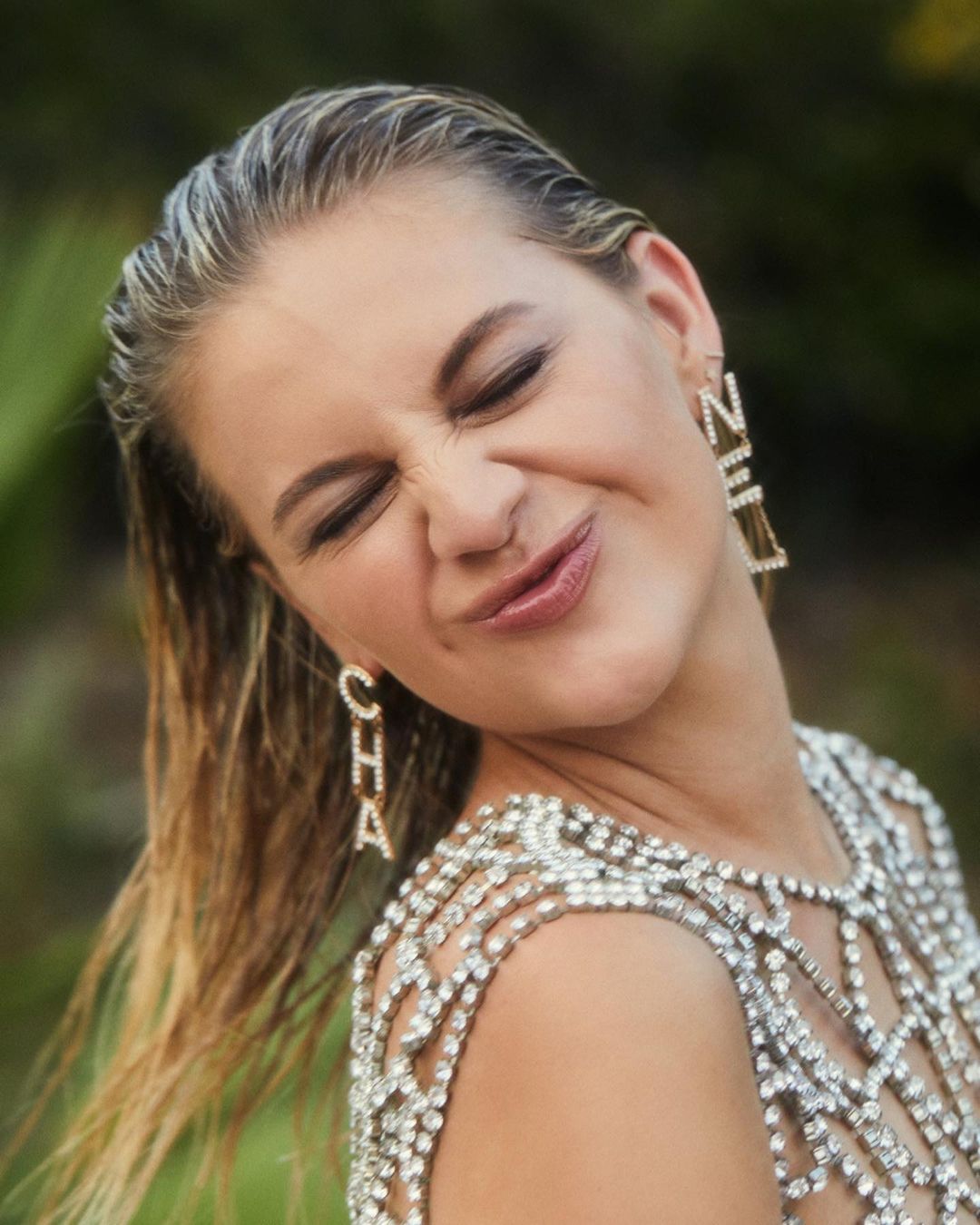 Close-up portrait of a blonde adult female squinting her eyes and sticking her tongue out, wearing sparkling dress and jewelry.