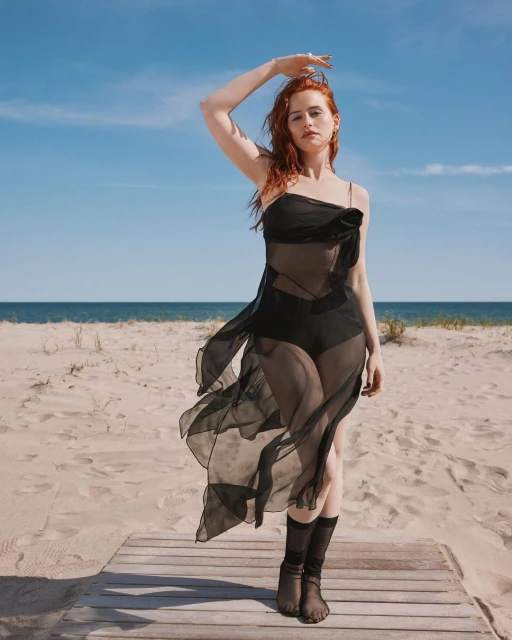 Red-haired woman in sheer black dress poses on a beach boardwalk under a blue sky with wispy clouds and ocean backdrop on a sunny day.