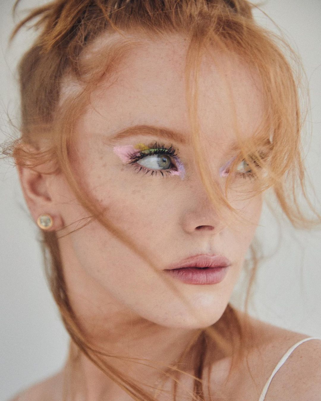 Close-up portrait of a red-haired woman with colorful eye makeup, fair skin, and an earring, against a neutral background in soft lighting.