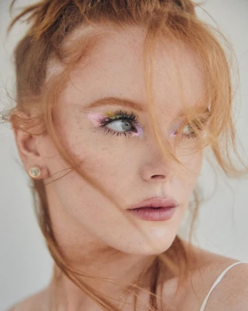 Close-up portrait of a red-haired woman with colorful eye makeup, fair skin, and an earring, against a neutral background in soft lighting.
