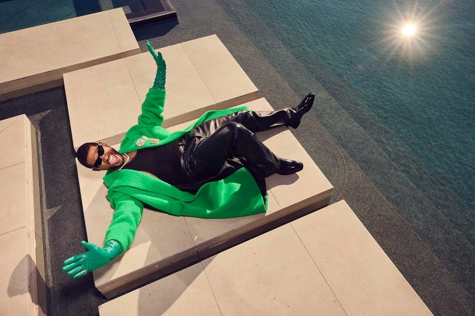 Usher poses in bright green coat, black pants and sunglasses by an outdoor pool on geometric tiles, arms spread.