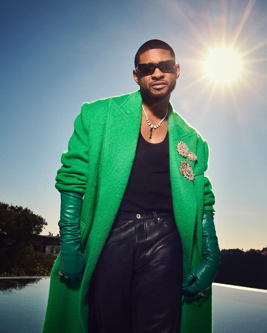 Usher wearing a green coat and sunglasses poses outdoors, with a pool and blue sky in the background, under bright sunlight.