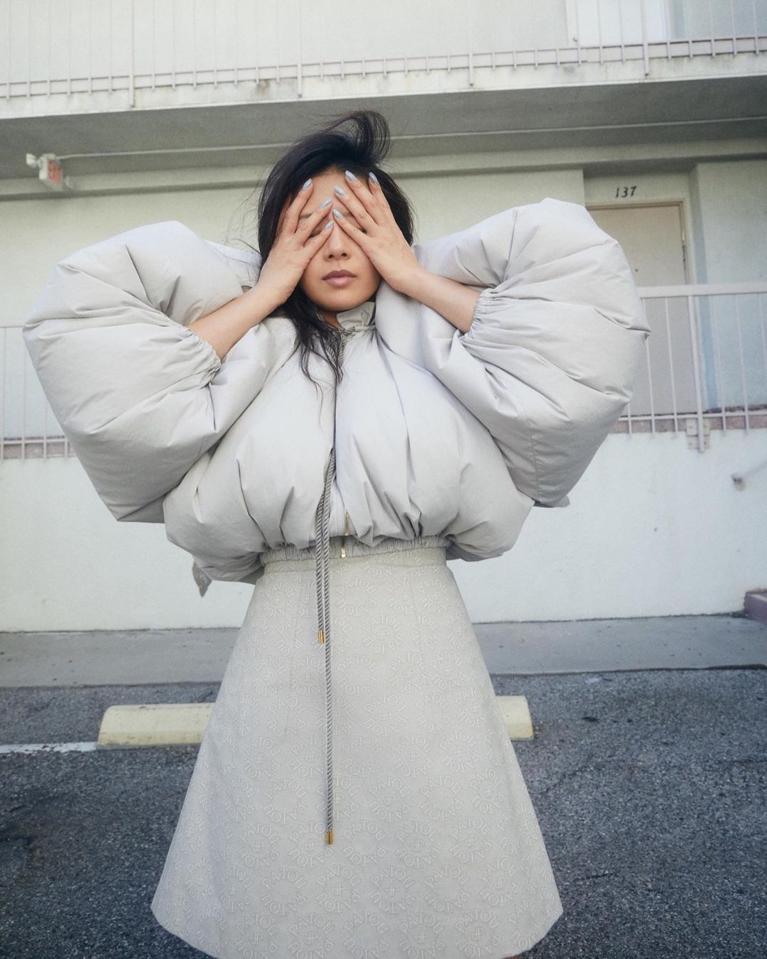 A woman in a padded jacket and skirt poses with her hands over her eyes, in an outdoor setting near a building with numbered doors.