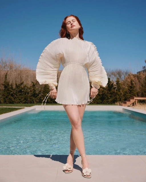Elegant red-haired woman posing in a short white pleated dress and flower-embellished heels, standing near a pool against a blue sky.