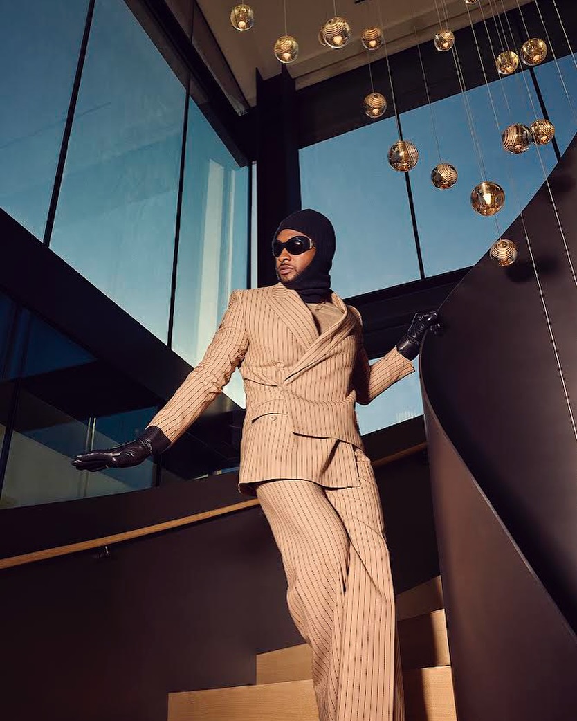 Stylish adult male in a pinstripe suit poses on a modern staircase with globe lights, wearing sunglasses, a beanie, and gloves.
