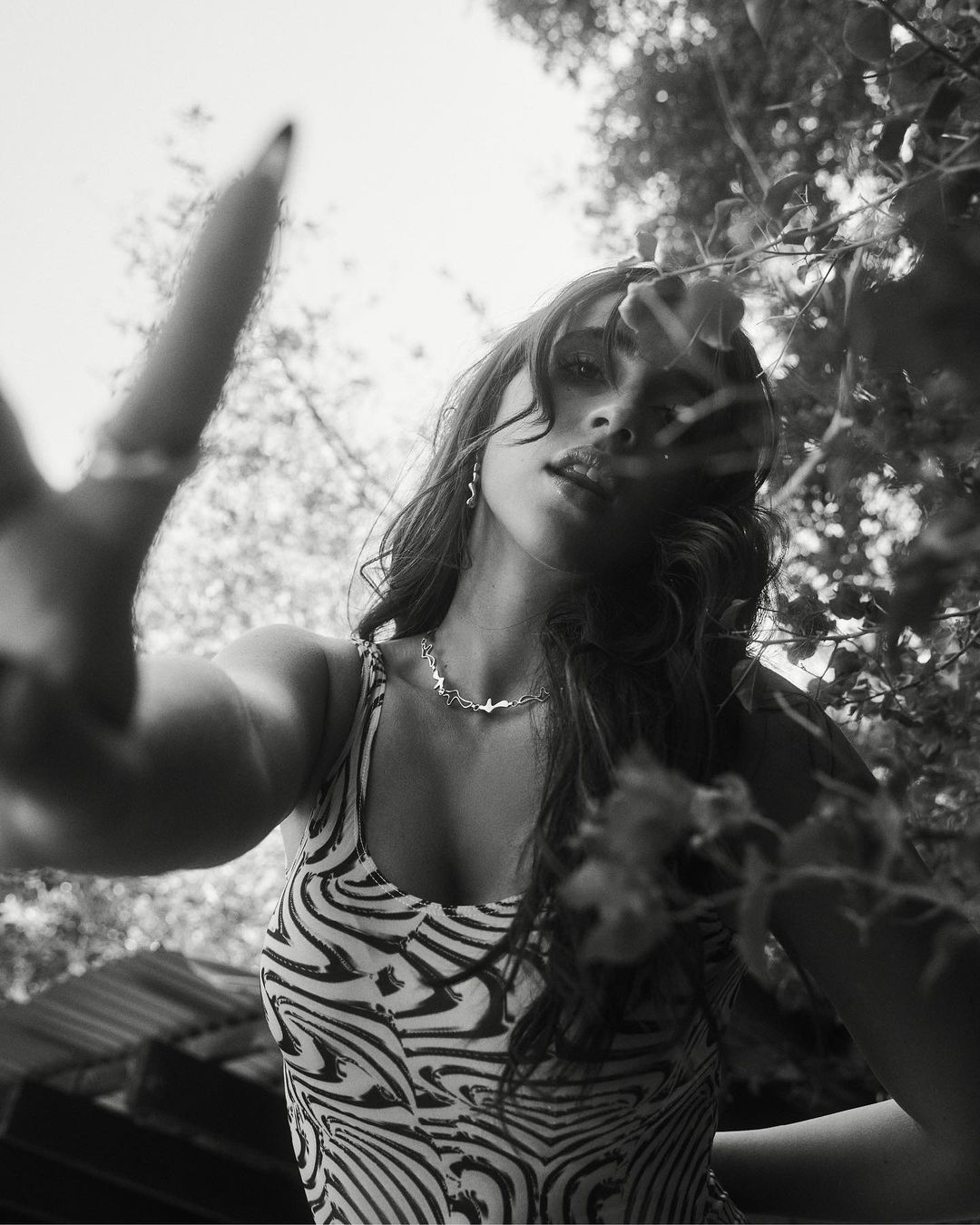 Low-angle view of a smiling woman in a zebra-stripe top, wearing a necklace, with leaves and sky visible in the background in black and white.