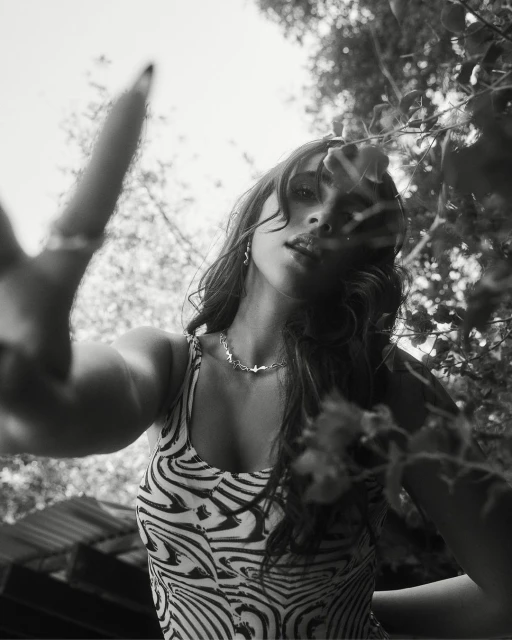 Low-angle view of a smiling woman in a zebra-stripe top, wearing a necklace, with leaves and sky visible in the background in black and white.