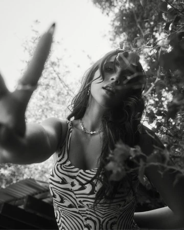 Low-angle view of a smiling woman in a zebra-stripe top, wearing a necklace, with leaves and sky visible in the background in black and white.