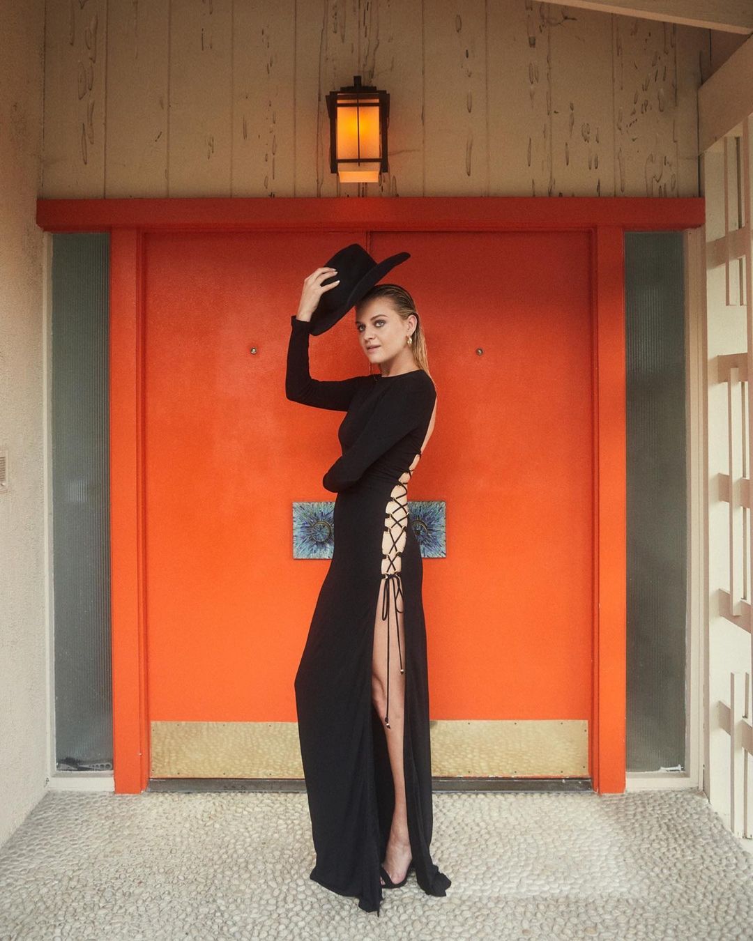 Elegant woman in a long black dress poses in front of a bright orange door, holding a black hat, showcasing fashion and style.