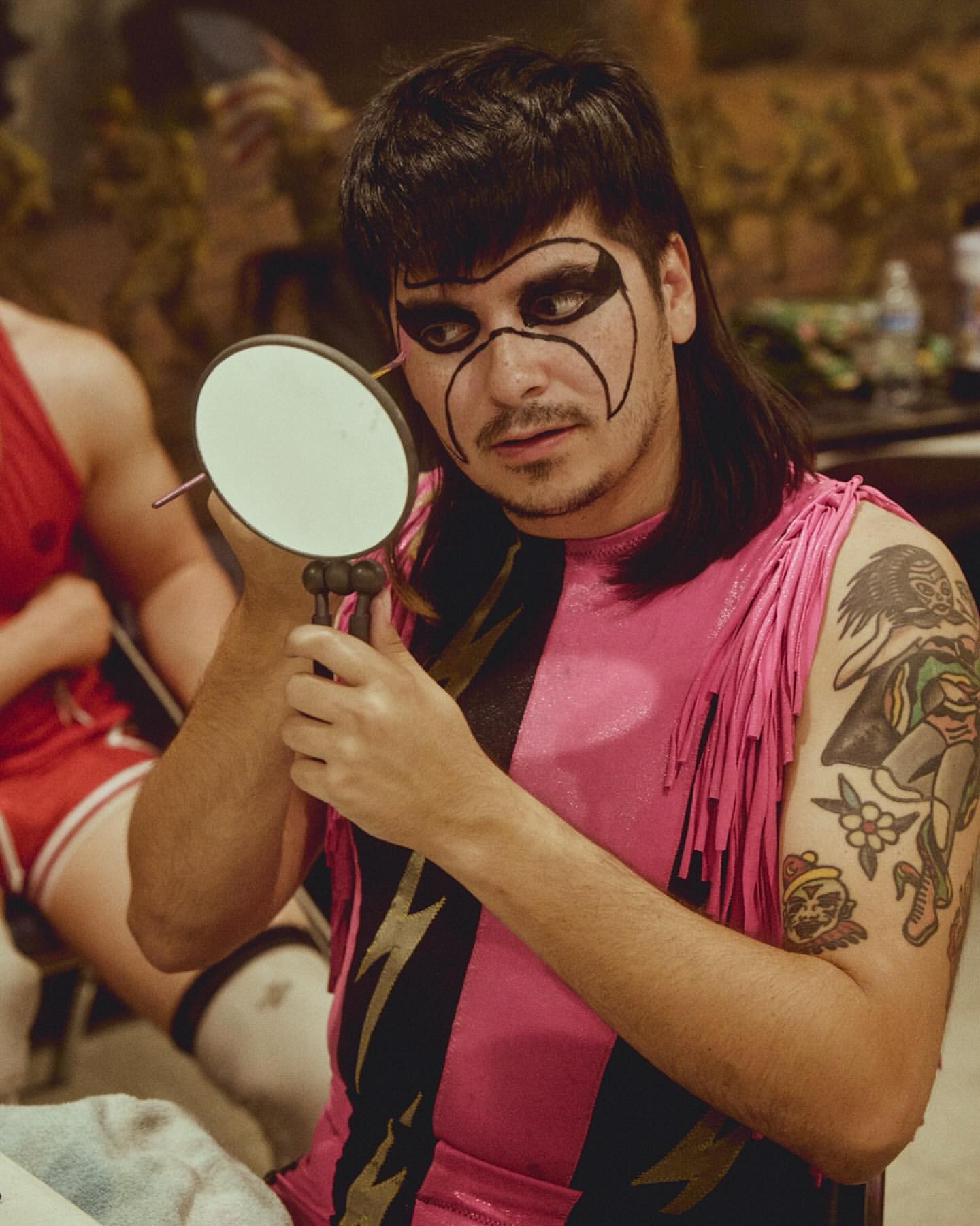 Adult male in pink and black wrestling suit with face paint applies makeup with a handheld mirror in an indoor dressing room.