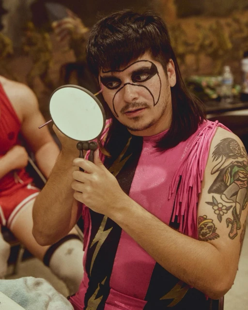 Adult male in pink and black wrestling suit with face paint applies makeup with a handheld mirror in an indoor dressing room.