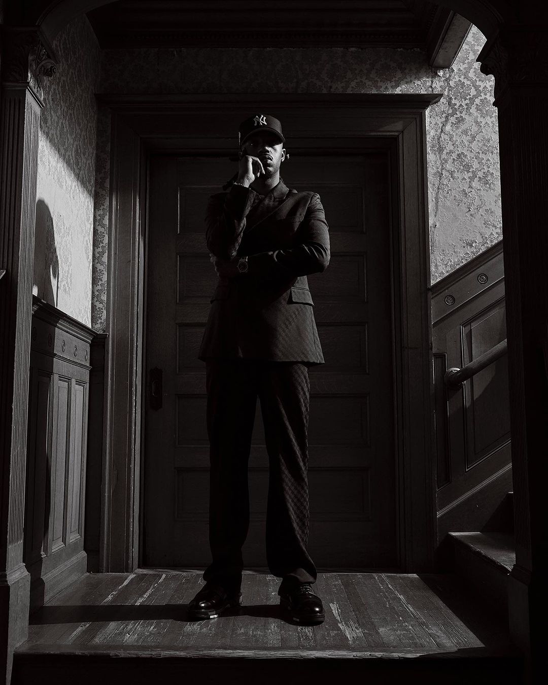 Man wearing a suit and baseball cap standing in a dimly lit hallway with patterned wallpaper
