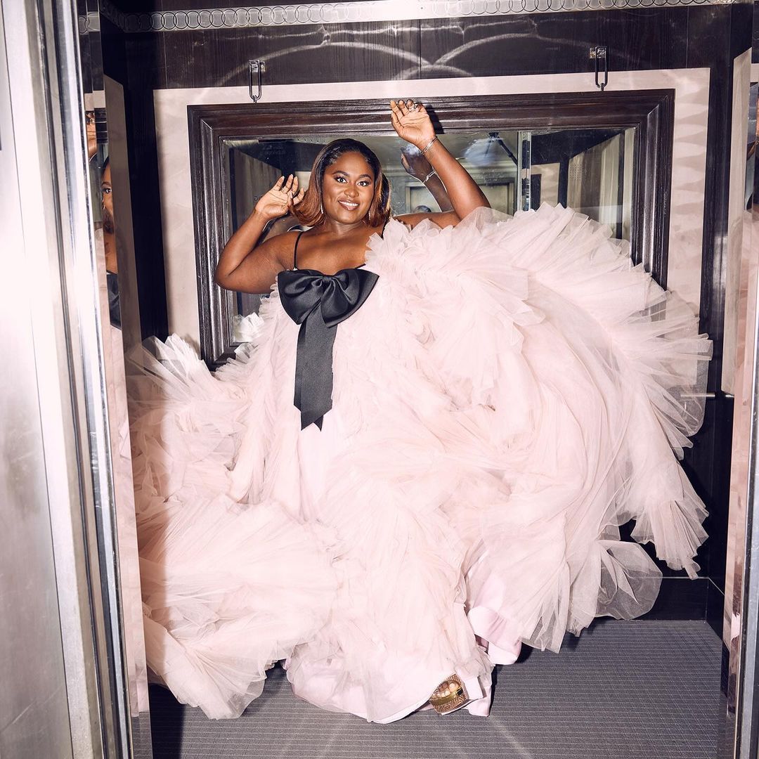 An African-American woman poses, smiling, in a light pink, tulle formal dress with a large black bow in an interior setting.