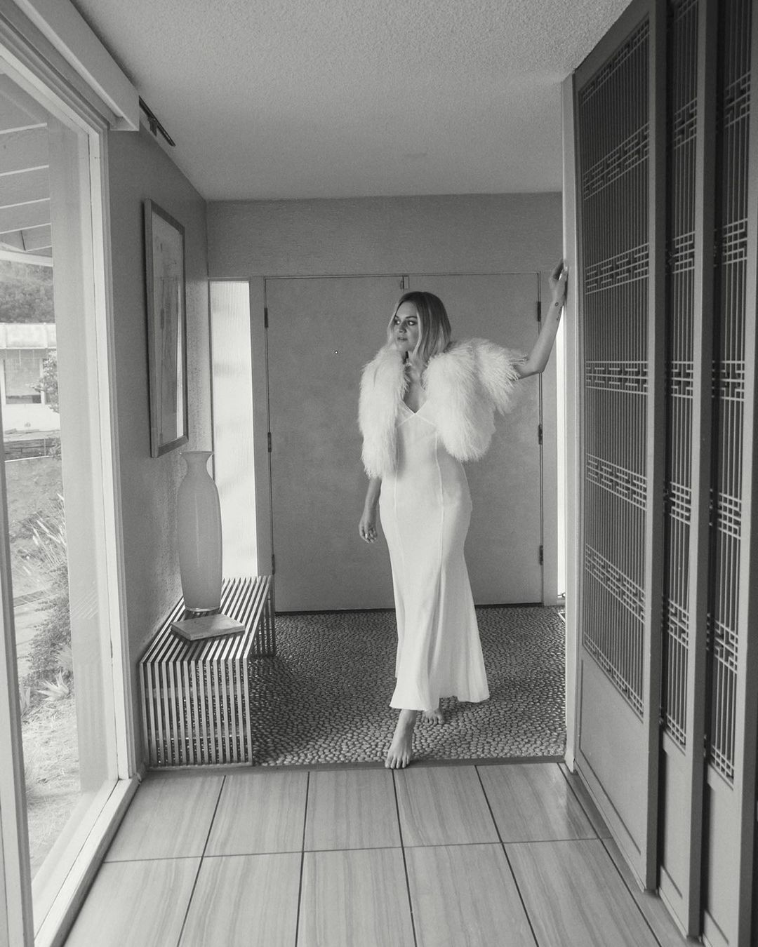 Monochrome portrait of an adult female in a white dress with a feathered bolero standing in an indoor hallway near a door.