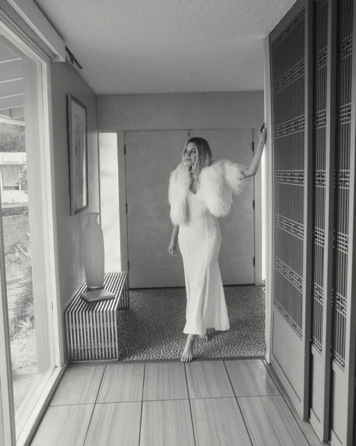 Monochrome portrait of an adult female in a white dress with a feathered bolero standing in an indoor hallway near a door.