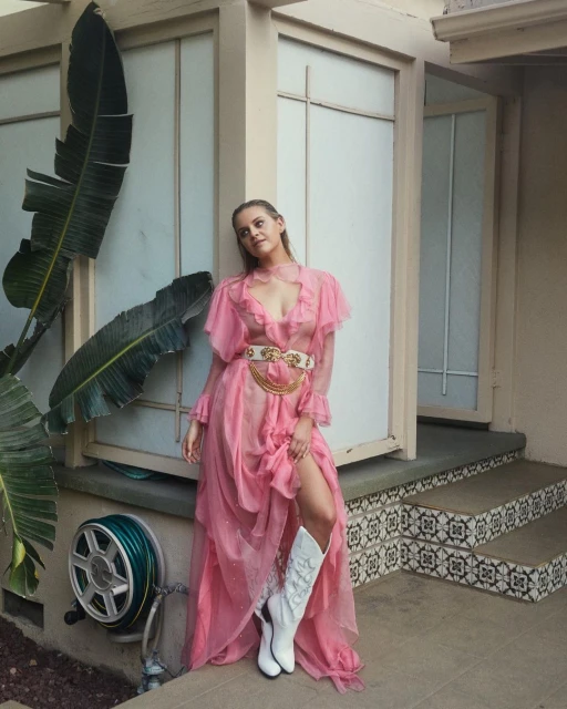 A young woman in a long pink dress and white cowboy boots poses in front of an exterior wall, with a large banana leaf plant nearby.