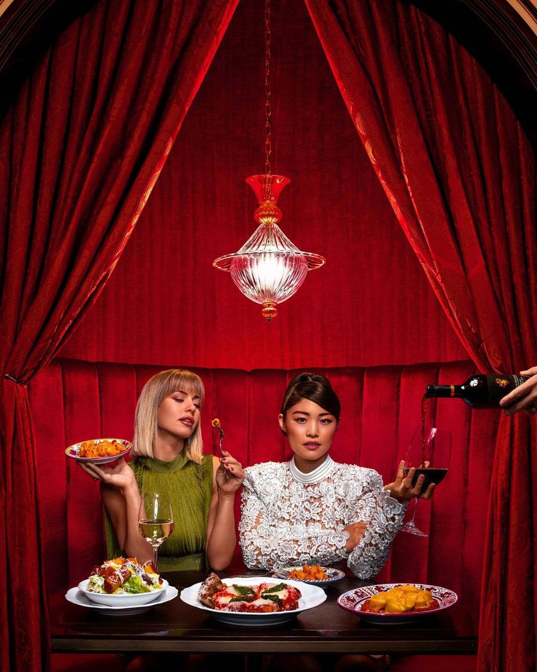 Two women seated at a restaurant booth with a table full of food, red curtains, and a hanging light fixture. Wine pouring into glass.