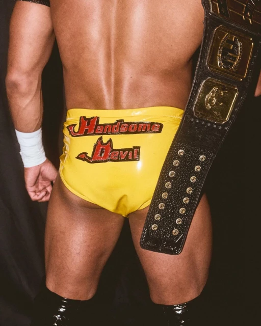 Close-up from the back of a muscular male wrestler wearing yellow trunks with the text 'Handsome Devil' and holding a championship belt.