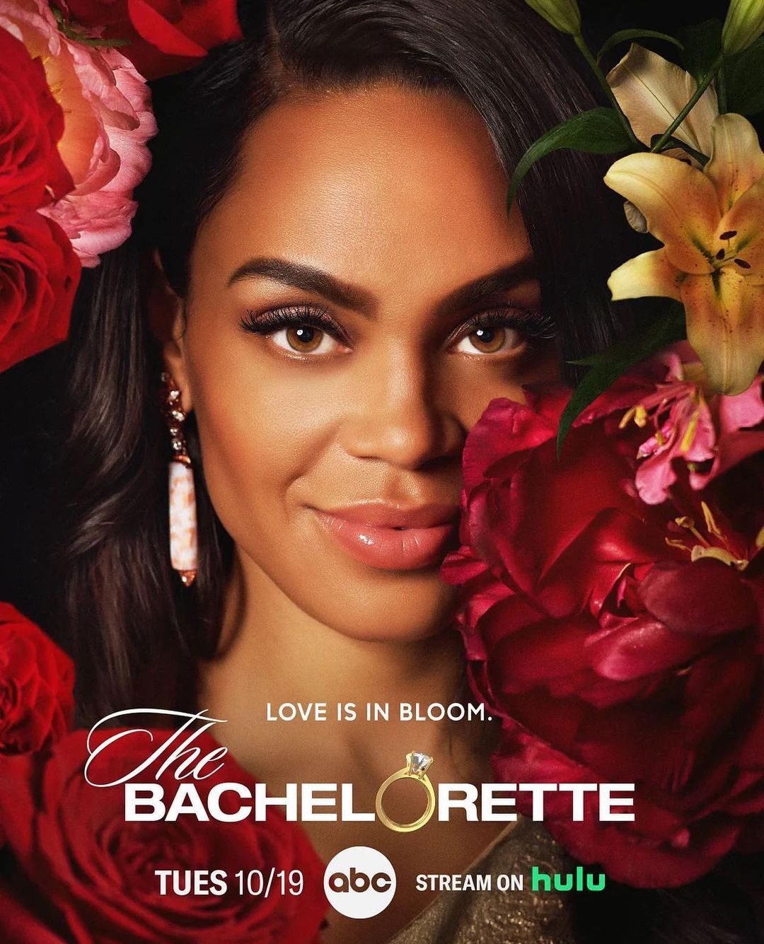 Close-up promotional image of a woman with tan skin and brown hair wearing earrings, posing amidst red and yellow flowers.
