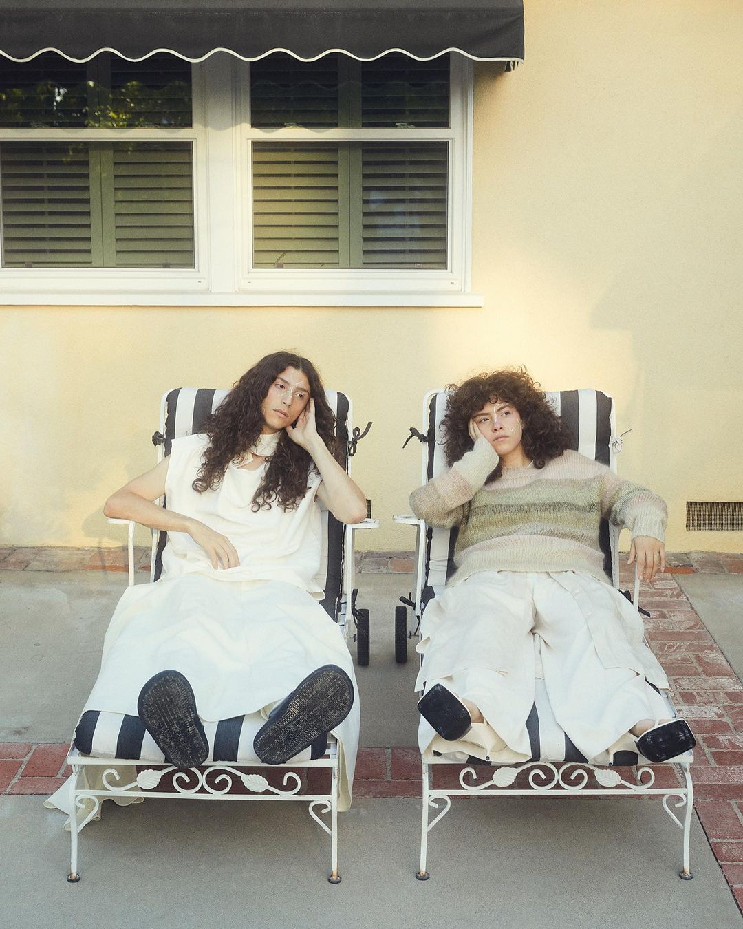 Two women sitting in striped lounge chairs outdoors, one in white dress, one in a striped sweater, against a tan wall in soft sunlight.