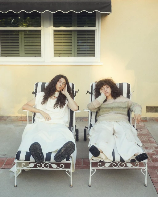 Two women sitting in striped lounge chairs outdoors, one in white dress, one in a striped sweater, against a tan wall in soft sunlight.