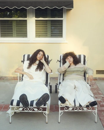 Two women sitting in striped lounge chairs outdoors, one in white dress, one in a striped sweater, against a tan wall in soft sunlight.