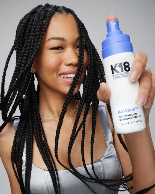 A smiling woman with braids holds up a bottle of K18 AirWash dry shampoo, showcasing haircare product and styling in a studio setting.