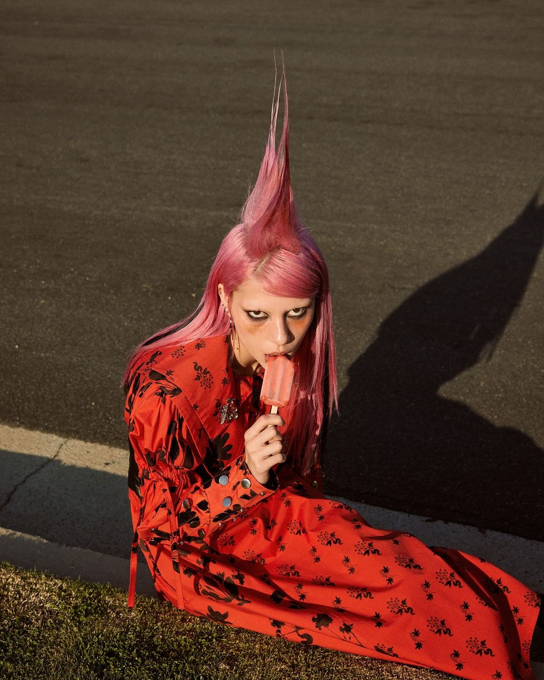 Adult female with pink hair and red floral dress, sitting on grass while eating an ice cream bar outside on a sunny day.